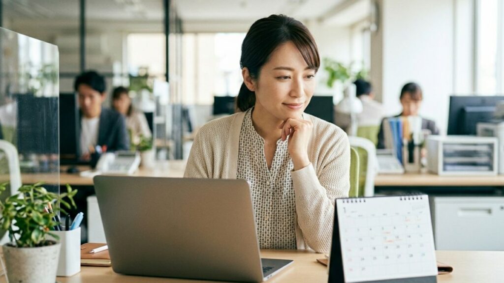 オフィスのデスクでカレンダーを眺めながら、有給休暇の計画を立てる笑顔の派遣社員の女性。